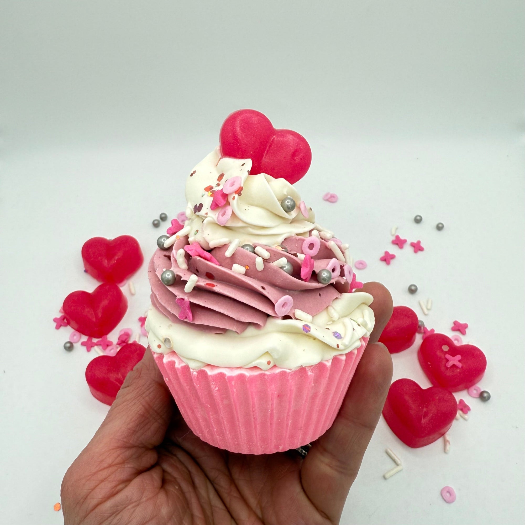 A handmade cupcake soap from Rusted Acre Soap Co. Pink cake topped with pink and white frosting and a pink heart soap embed. Held in a hand.