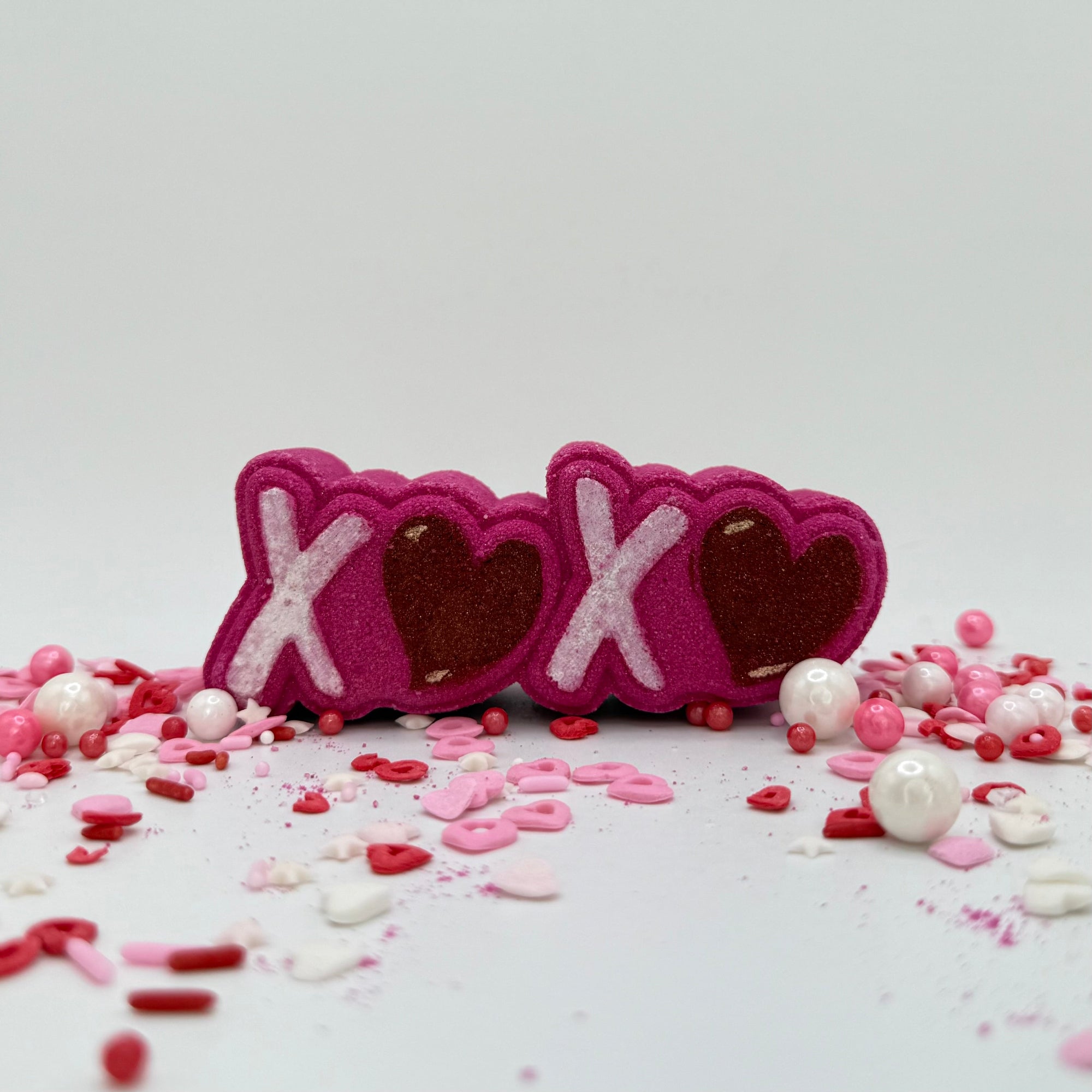 Pink bath bomb with raised white X's and pink hearts. Displayed with pink sprinkles of various shapes.